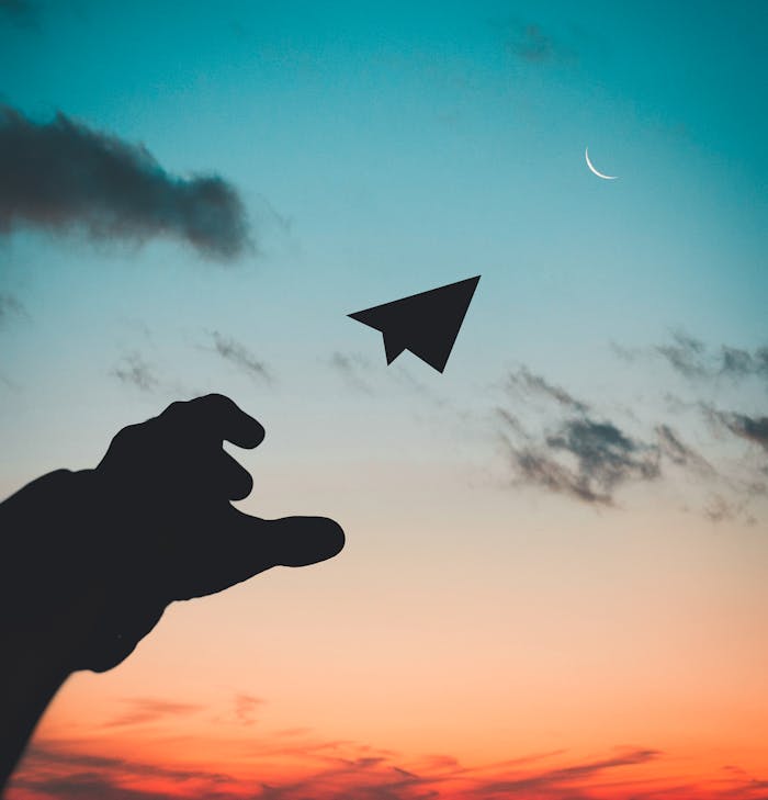 about-us-01 A silhouette of a hand launching a paper plane against a colorful dusk sky with a crescent moon.